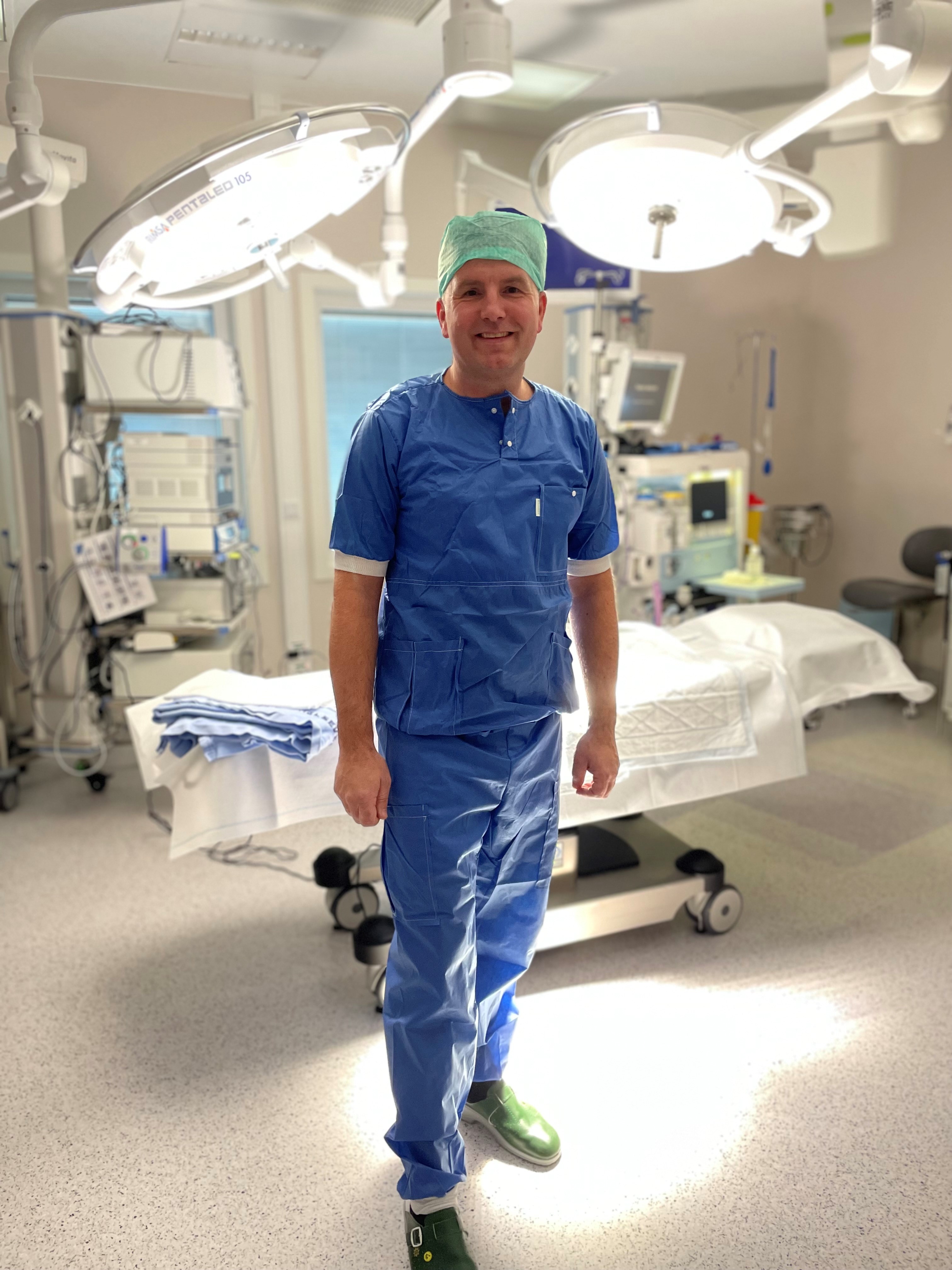 Mann i operasjonsklær fra operasjonsstue smilernde mot  kamera