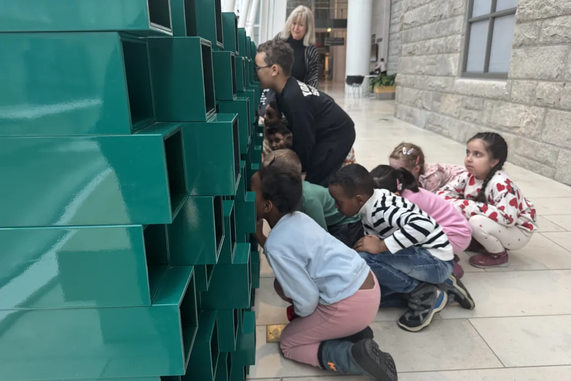 En gruppe barn som sitter på bakken ved siden av en grønn skulptur