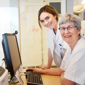 To damer ved PC som ser mot kamera og smiler